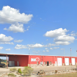 A view of the Labelle Volunteer Fire Department - Station 1, taken from the roadway. The facility is a large red metal building. The front of the building has been adorned with stone work for the first few feet. The North end of the building is visible in the left part of the image. It consists of a large covered area that is open on three sids and has a massive grill/smoker under the edge of it. The middle part of the building is administrative and community area, with a few windows, a glass door, another window and a white door. The remainder of the building is three floor to roof white roll-up doors, each with a flag pole on the right side of the door ( one between each of the doors, and a third after the last door). Rocks and vegetation is visible in the foreground with a beautiful blue sky and large fluffy clouds lazily passing of into the distance.