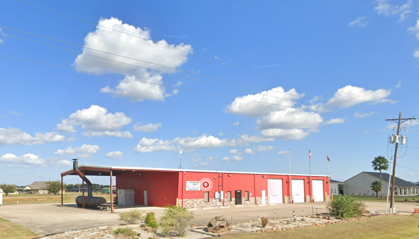 A view of the Labelle Volunteer Fire Department - Station 1, taken from the roadway. The facility is a large red metal building. The front of the building has been adorned with stone work for the first few feet. The North end of the building is visible in the left part of the image. It consists of a large covered area that is open on three sids and has a massive grill/smoker under the edge of it. The middle part of the building is administrative and community area, with a few windows, a glass door, another window and a white door. The remainder of the building is three floor to roof white roll-up doors, each with a flag pole on the right side of the door ( one between each of the doors, and a third after the last door). Rocks and vegetation is visible in the foreground with a beautiful blue sky and large fluffy clouds lazily passing of into the distance.