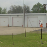 A photo of the Fannett Volunteer Fire Department - Station 2 is shown from the roadway. The building is a white metal construction and is older with three equally spaced floor to roof roll-up doors. Two fire hydrants separate the doors. A sign is visible on the side of the building, just above head high. It is not legible from this viewpoint. A large concrete pad is in front of the building .