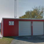 An image captured from the roadway of the front of the Cheek Fire Station - Station #3 on Kidd Road. The building is red with white trim and has two floor to roof white roll-up doors on the right side of the building. The left side of the building has a white CHEEK VOLUNTEER FIRE DEPARTMENT sign displayed. An antennae can be seen rising from the rear of the building and a fire engine is parked in the back of the lot facing the side gate.